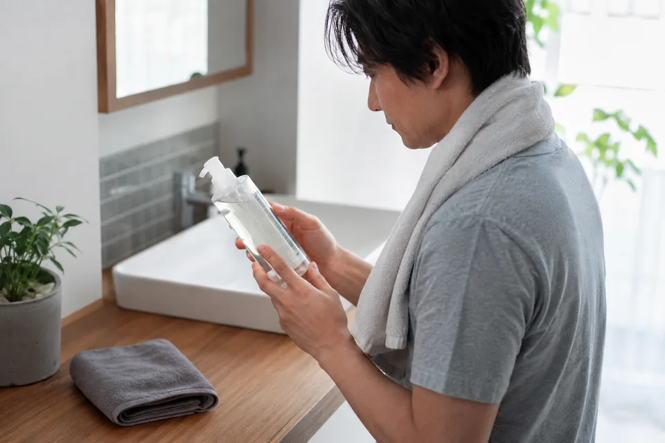 洗面台の前で、透明なポンプ容器の裏面の表示を読む男性